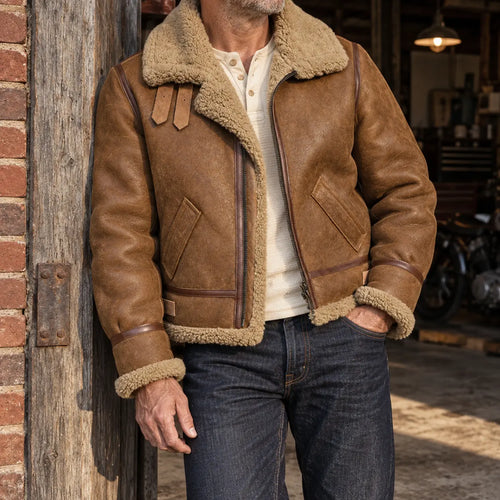 A man wearing a brown leather aviator-style jacket with a thick beige shearling lining, styled with a cream henley shirt and dark blue jeans, leaning against a rustic wooden post in a garage setting.